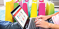 Frau sitzt auf Teppich mit Laptop zum Online-Shopping neben bunten Einkaufstaschen