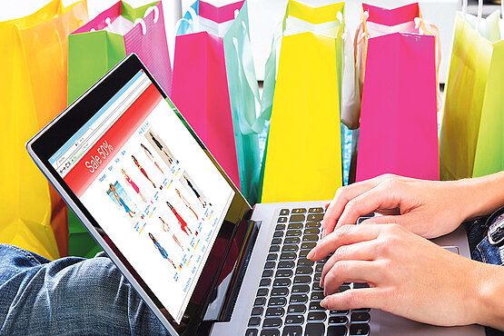 Frau sitzt auf Teppich mit Laptop zum Online-Shopping neben bunten Einkaufstaschen