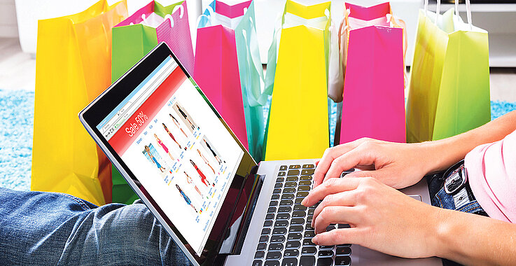Frau sitzt auf Teppich mit Laptop zum Online-Shopping neben bunten Einkaufstaschen