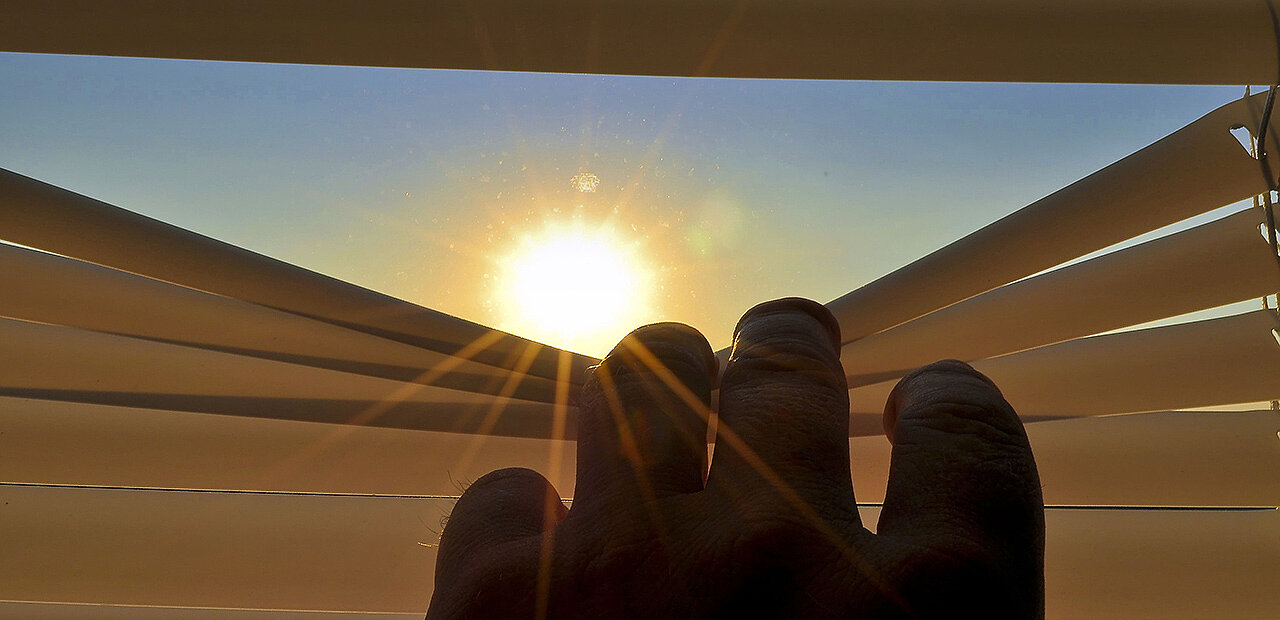 Ein Hand zieht ein Jalousie zur seite und gibt damit die Sicht auf die Sonne frei.