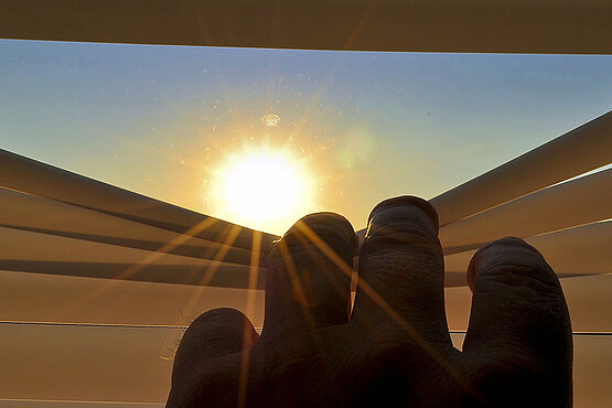 Ein Hand zieht ein Jalousie zur seite und gibt damit die Sicht auf die Sonne frei.