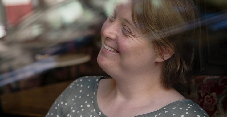 Eine Frau mit Down-Syndrom sitzt hinter einer Glasscheibe.