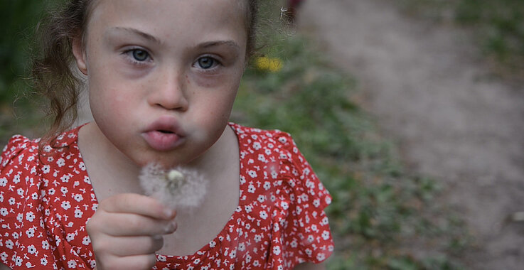 Ein Mädchen mit Down-Syndrom bläßt auf eine Pusteblume.