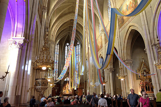 Pfingstinstallation Dein Atem – ruach Elohim von Franziska Pirstinger in der Herz-Jesu-Kirche in Graz. Wie ein großer Lichtstrahl fallen sieben Farbbahnen aus dem Heiliggeistloch. Die sieben Gaben des Geistes breiten sich wie ein Zelt über den Gläubigen aus. Der Atem Gottes heilt und erneuert Kirche und Welt.