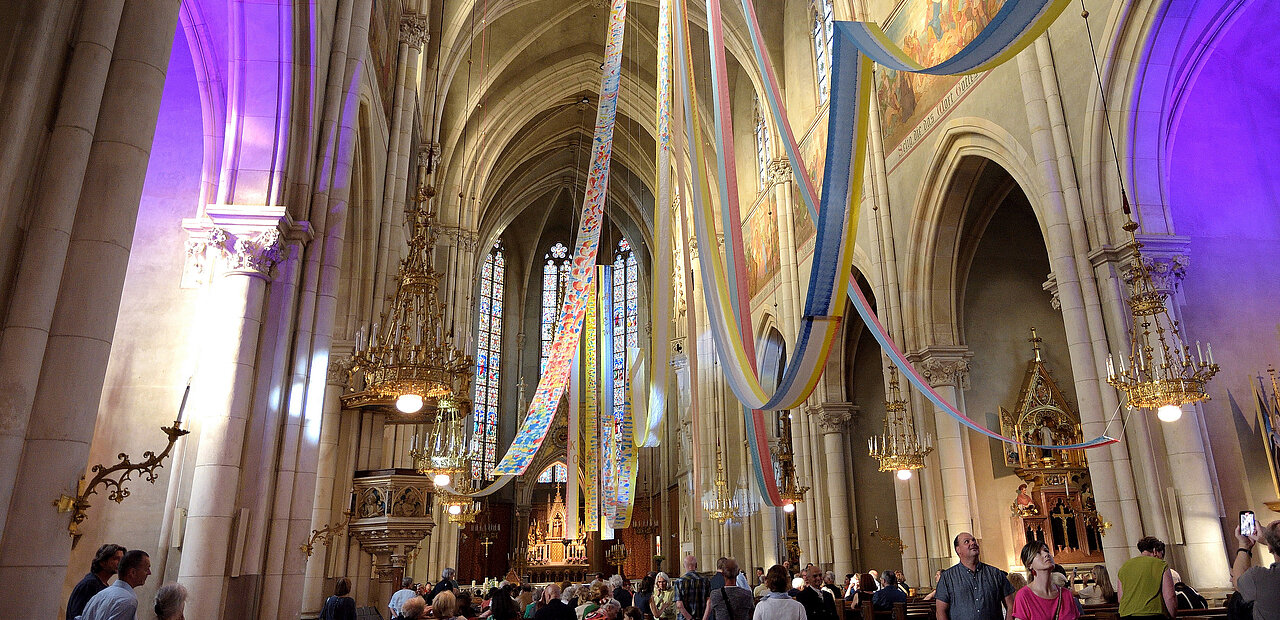 Pfingstinstallation Dein Atem – ruach Elohim von Franziska Pirstinger in der Herz-Jesu-Kirche in Graz. Wie ein großer Lichtstrahl fallen sieben Farbbahnen aus dem Heiliggeistloch. Die sieben Gaben des Geistes breiten sich wie ein Zelt über den Gläubigen aus. Der Atem Gottes heilt und erneuert Kirche und Welt.