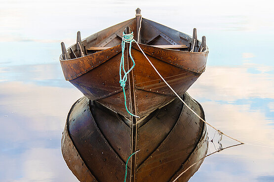 Ein kleines Holzruderboot, dass sich im Stillen Wasser spiegelt.