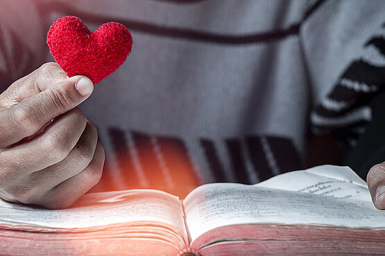 Jemand hält ein rotes Herz in der rechten Hand und liest dabei in der Bibel.