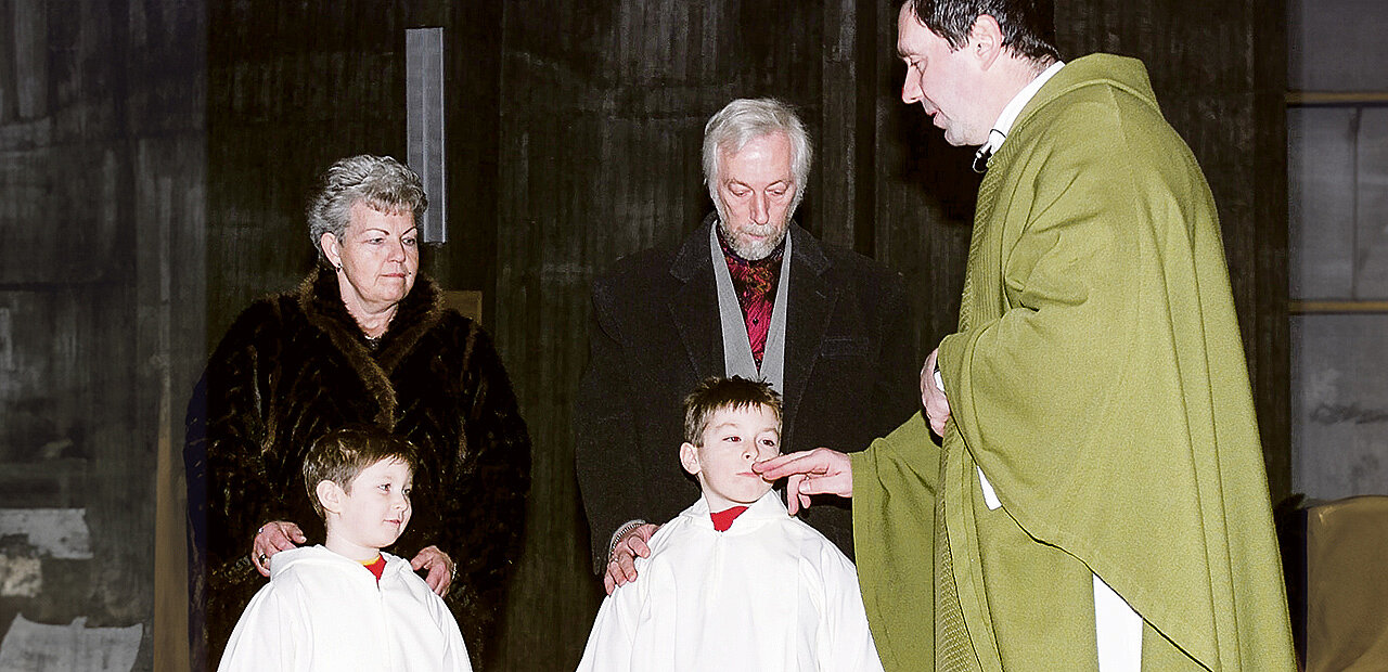 Zwei Kinder werden getauft. Am Bild zu sehen die Kinder, und deren Großeltern und der Priester Der Priester berührt den Mund eines der Täuflinge.