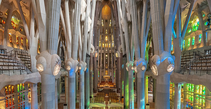 Sakraler Wald: Blick durch das Hauptschiff auf den Altar der Sagrada Família, getragen von sich verzweigenden Säulen und erfüllt von farbig schimmerndem Licht.